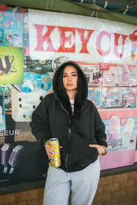 Oversized cropped pigment black hoodie by Alphabet City, heavyweight 500+ GSM 100% cotton with terry inside, double silver A logo zip pullers, drop shoulder fit, cord drawstring with stoppers, and embroidered A logo on sleeve. Unisex design, custom oversized cropped fit, shown on model Jassy wearing size medium. Free All Stars Miami T-shirt included with every order.